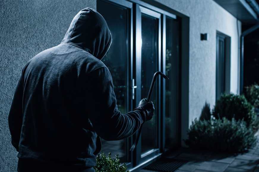 Dark figure wearing a hooded jacket attempts to break into a home at night by using a crowbar on a glass door to gain unauthorized access to the
