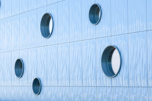 Kreisrunde Fenster aus GENEO Profilen setzen in der blauen Wellenfassade ansprechende Designakzente. - © REAL Fenster und Türen GmbH
