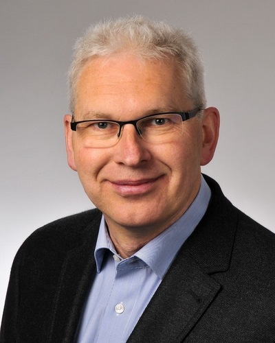Lächelnder Mann mit kurzen grauen Haaren, Brille und einem dunklen Blazer über einem blauen Hemd, vor einem schlichten Hintergrund. - © Frank Eßmann