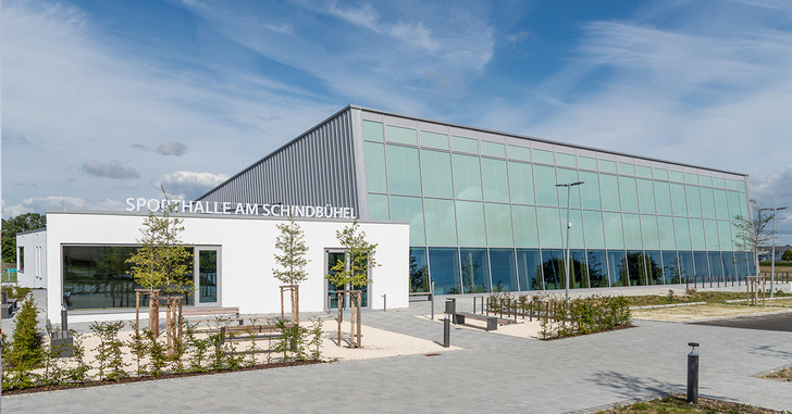 Moderne Sporthalle mit großen Glasfenstern, umgeben von Bäumen und einem gepflasterten Gehweg unter einem teilweise bewölkten Himmel. - © TA, Architektur: mvm+starke, Architektur: bharchitekten