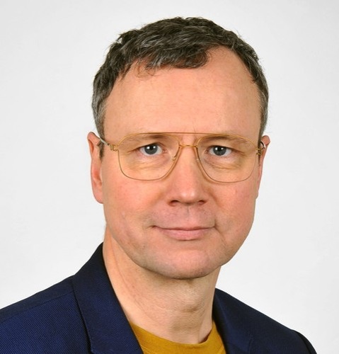 Mann mit kurzen braunen Haaren, der eine goldene Brille, einen blauen Blazer und ein gelbes Hemd trägt, vor einem schlichten Hintergrund. - © Oliver Solcher