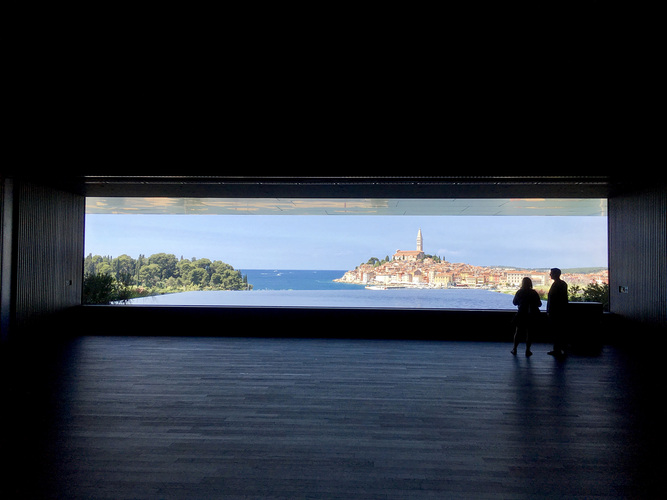 Für die Planer des neuen 5-Sterne-Hotels war früh klar, dass dem besonderen Ausblick die entscheidende Rolle in der Gestaltung zukommt. So wurden alle Räume mit freier Sicht auf Bucht und Stadt entworfen. Für den großen Ballsaal — als Herz des Hotels — hatten sie etwas Besonderes im Sinn: “Wir haben bei sedak angefragt, was in diesem Umfeld maximal möglich ist. Mit dieser Information haben wir den Saal geplant — sowohl sein Aussehen als auch die Position“, berichten die Architekten. Das Ergebnis war ein Isolierglas vom Format 3,2 x 15 m. Dieses XXL-Glas ist aus zwei 2-fach-Laminaten aufgebaut, die wiederum aus 2 x 10mm sowie 2 x 12mm TVG ausgebaut sind. “Ein so großes Glas lässt die Grenzen zwischen Innen und Außen verschwinden. Wir können damit atemberaubende, fast unreale Effekte erzeugen“, betonen die Architekten, ein Ergebnis das den Aufwand rechtfertigt. Ein XXL-Glas aus Gersthofen Damit die Effekte wie gewünscht gelingen, muss das Glas höchsten Qualitäts-Ansprüchen gerecht werden. In der sedak-Produktion wurde das 3,2 × 15 m Isolierglas vollautomatisch gefertigt. Gut verpackt erreichte das 5t schwere Glas Rovinj. Dort wurde es von einem Kran mit einem 50-m-Ausleger behutsam über das Hotel an seinen Bestimmungsort gehoben. Mit Hilfe von Führungsschienen wurde das makellose Glas in seine aufwendig geplante Halterung gebracht. Diese trägt neben der Scheibe noch einen dem Glas vorgelagerten, 5 m auskragenden Balkon, der das Fenster in Schatten hüllt. hüllt. Dadurch verstärkt sich die Transparenz. Zugleich liegt zu Füßen des Ballsaals eine große Wasserfläche, die die Reflexion der Altstadt auch beim Blick von Innen aufnimmt und an den Betrachter weitergibt. www.sedak.com Atemberaubender Blick aus dem Ballsaal auf die malerische Stadt und das Meer. Eine 15m lange Scheibe von sedak macht es möglich. - © sedak GmbH
Für die Planer des neuen 5-Sterne-Hotels war früh klar, dass dem besonderen Ausblick die entscheidende Rolle in der Gestaltung zukommt. So wurden alle Räume mit freier Sicht auf Bucht und Stadt entworfen. Für den großen Ballsaal — als Herz des Hotels — hatten sie etwas Besonderes im Sinn: “Wir haben bei sedak angefragt, was in diesem Umfeld maximal möglich ist. Mit dieser Information haben wir den Saal geplant — sowohl sein Aussehen als auch die Position“, berichten die Architekten. Das Ergebnis war ein Isolierglas vom Format 3,2 x 15 m. Dieses XXL-Glas ist aus zwei 2-fach-Laminaten aufgebaut, die wiederum aus 2 x 10mm sowie 2 x 12mm TVG ausgebaut sind. “Ein so großes Glas lässt die Grenzen zwischen Innen und Außen verschwinden. Wir können damit atemberaubende, fast unreale Effekte erzeugen“, betonen die Architekten, ein Ergebnis das den Aufwand rechtfertigt. Ein XXL-Glas aus Gersthofen Damit die Effekte wie gewünscht gelingen, muss das Glas höchsten Qualitäts-Ansprüchen gerecht werden. In der sedak-Produktion wurde das 3,2 × 15 m Isolierglas vollautomatisch gefertigt. Gut verpackt erreichte das 5t schwere Glas Rovinj. Dort wurde es von einem Kran mit einem 50-m-Ausleger behutsam über das Hotel an seinen Bestimmungsort gehoben. Mit Hilfe von Führungsschienen wurde das makellose Glas in seine aufwendig geplante Halterung gebracht. Diese trägt neben der Scheibe noch einen dem Glas vorgelagerten, 5 m auskragenden Balkon, der das Fenster in Schatten hüllt. hüllt. Dadurch verstärkt sich die Transparenz. Zugleich liegt zu Füßen des Ballsaals eine große Wasserfläche, die die Reflexion der Altstadt auch beim Blick von Innen aufnimmt und an den Betrachter weitergibt. www.sedak.com Atemberaubender Blick aus dem Ballsaal auf die malerische Stadt und das Meer. Eine 15m lange Scheibe von sedak macht es möglich. - sedak GmbH