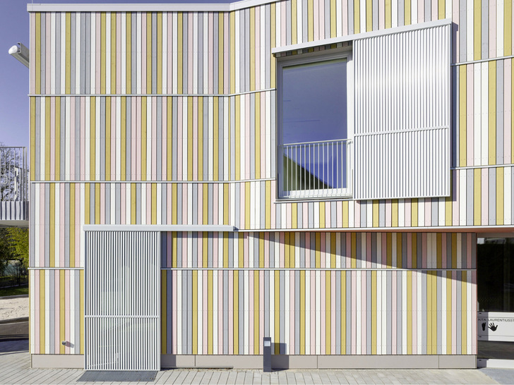  Die vertikalen Lamellen passen zur aufstrebenden Ausrichtung der bunten Fassadeleisten; das Grau der Schiebeläden und Blenden wiederholt sich ebenfalls in der gestrichenen Holzfassade und taucht sogar in den horizontalen Fassadenleisten auf. 