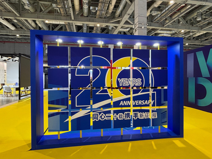 Optimierter Alt-Text: „Digitaler Messestand mit Jubiläumsanzeige „20 Jahre“ auf großem blauen Display, gelbe Akzente und industrielle Decke; Szene repräsentiert moderne Präsentation von Fenster- und Fassadentechnik für die Bauelemente-Branche.“.