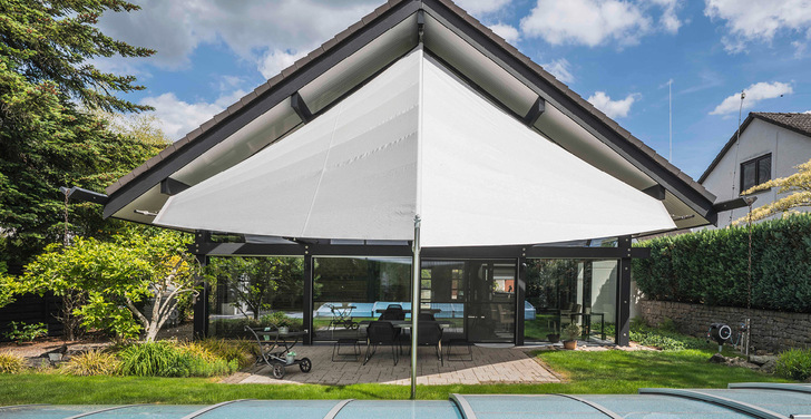 Vollflächig beschattet – technisch anspruchsvoll umgesetzt: Das markante Doppeldreiecksegel mit 7 m Wellen­länge und 5,5 m Auszug beschattet nun die gesamte Hausfront inklusive Terrasse. Die Montage am Dachfirst stellte durch die Höhe hohe Anforderungen an Befestigung und Einbringung der Tuchwelle.