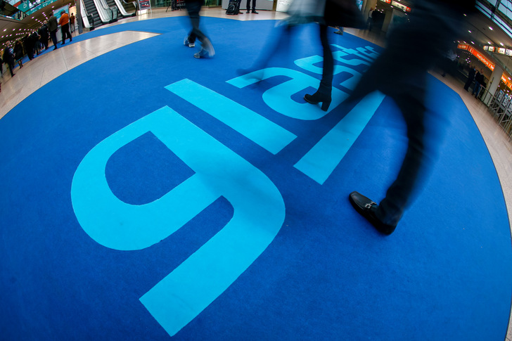 Menschen laufen über einen blauen Boden, auf dem in großen hellblauen Buchstaben "glasstec" steht. - © Messe Düsseldorf / ctillmann