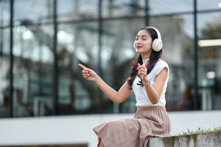 Frau, die draußen sitzt, Kopfhörer trägt, lächelt und mit den Fingern zeigt, während sie Musik genießt. - © Saint-Gobain Glass