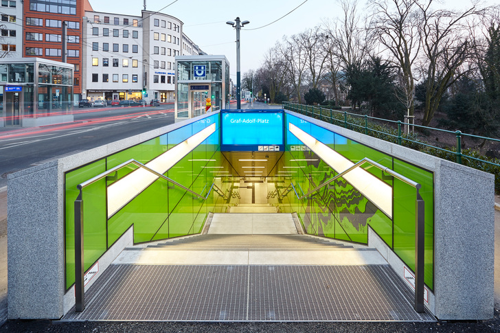 Dieser U-Bahn-Halt in Düsseldorf ist ab den Zugängen mit Glas gestaltet. Auf den rechten Glastafeln sieht man bereits den sogenannten "Linienstrom", der die Fahrgäste bis zum Bahnsteig hinab begleitet.