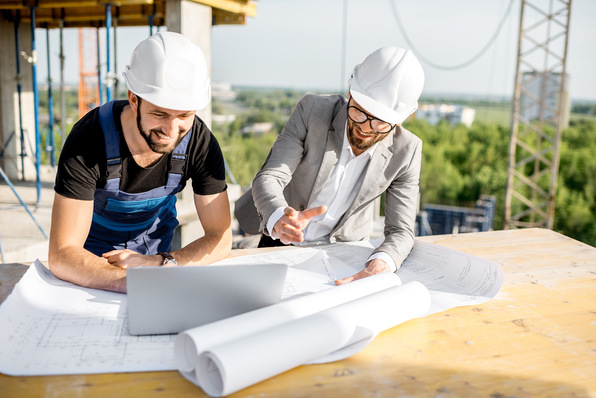 Engineer with worker in uniform working with architectural drawings and laptop at the table on the construction site outdoors