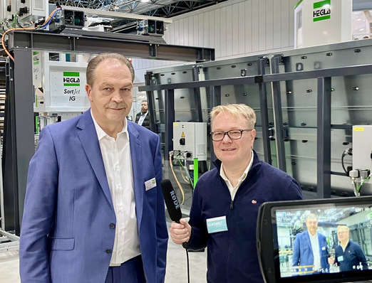 Alt-Text: „Zwei Fachleute diskutieren auf einer Fachmesse über innovative Fenstersysteme, umgeben von moderner Fassadentechnologie. - © GLASWELT