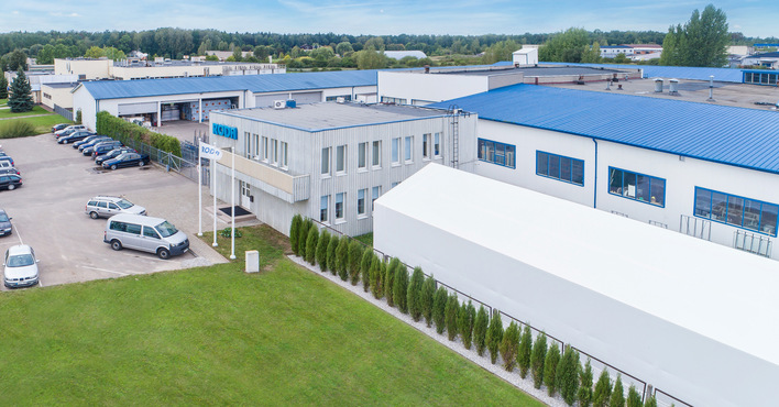 Sicht auf ein modernes Industriegebäude mit blauen Dächern, strategisch platziertem Parkplatz und weißer Transporterflotte. Im Vordergrund sind Flaggenmasten und eine gepflegte Grünanlage sichtbar, was die nachhaltige Integration in die umgebende Landschaft unterstreicht. Das Bild repräsentiert innovative Fassadensysteme und bautechnische Effizienz innerhalb der Glas- und Bauelementebranche. - © Roda