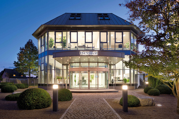 Modern glass building with illuminated "heroal" sign, surrounded by trees and shrubs in evening light. - © heroal