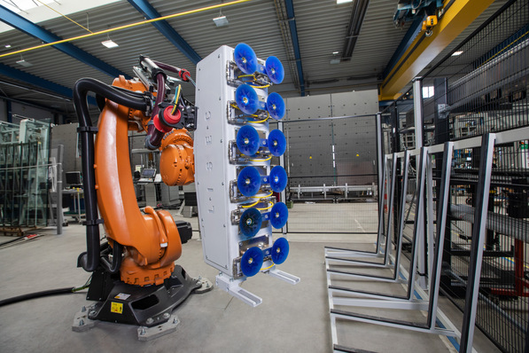„Industrieroboter mit Saugnäpfen bei der Montage von Fensterrahmen in einer Produktionsanlage, umgeben von Aluminiumprofilen und Isolierglaseinheiten. - © Lisec