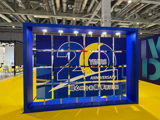 Optimierter Alt-Text: „Digitaler Messestand mit Jubiläumsanzeige „20 Jahre“ auf großem blauen Display, gelbe Akzente und industrielle Decke; Szene repräsentiert moderne Präsentation von Fenster- und Fassadentechnik für die Bauelemente-Branche.“.