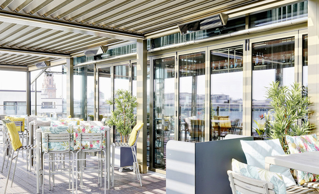 Die vierteilige Anlage mit dem Lamaxa L50 fügt sich mit der Sonder-Pulverbeschichtung im Farbton Mittelbronze harmonisch in die Architektur der Dachterrasse ein und verbindet Design, Komfort sowie flexiblen Wetterschutz.
