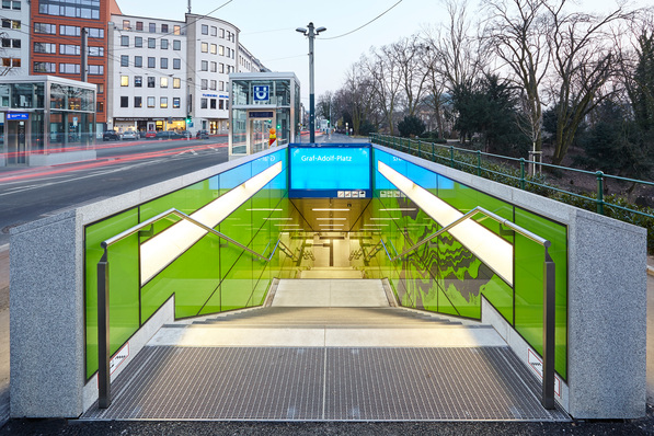 Dieser U-Bahn-Halt in Düsseldorf ist ab den Zugängen mit Glas gestaltet. Auf den rechten Glastafeln sieht man bereits den sogenannten "Linienstrom", der die Fahrgäste bis zum Bahnsteig hinab begleitet.